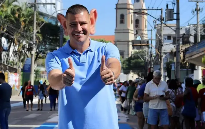 Montagem: político sorrindo e fazendo joinha na rua, com orelhas gigantes.